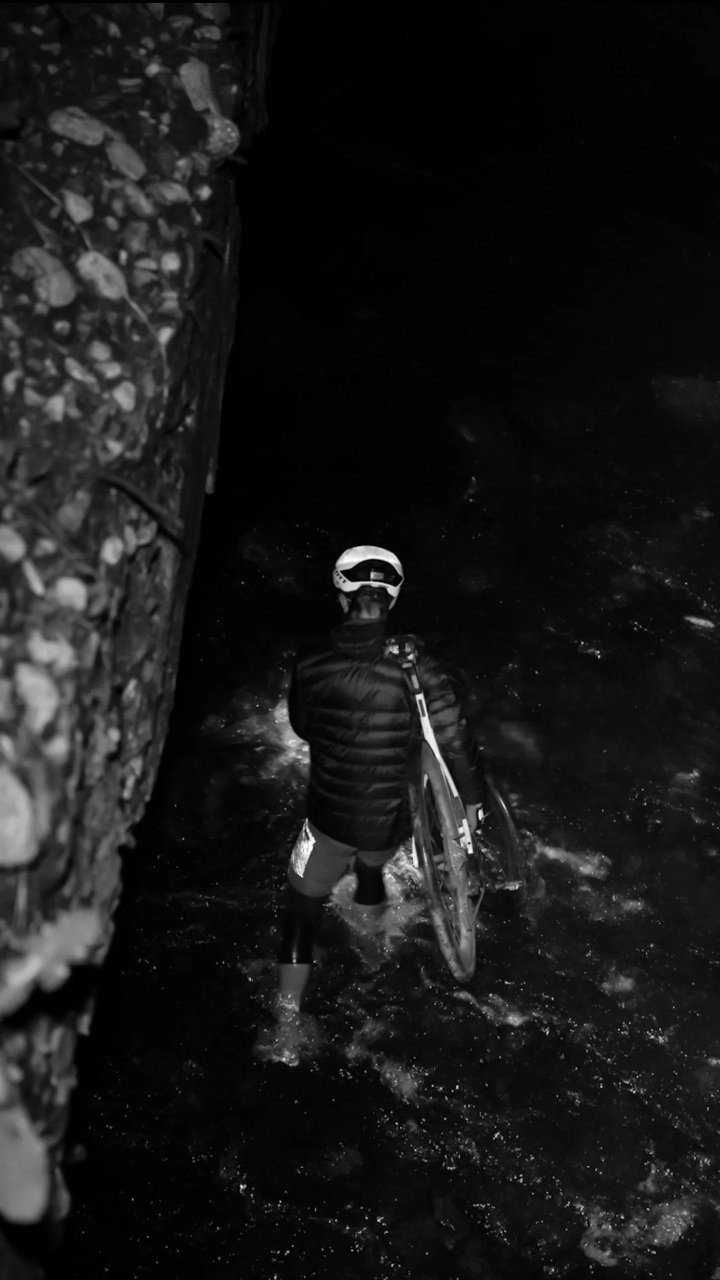Eine Person in einer schwarzen Jacke und mit Helm steht nachts im Wasser und hält ein Fahrrad in der Hand. Die Szene ist dunkel, links ist ein felsiger Rand zu sehen. Schwarz-Weiß fotografiert.