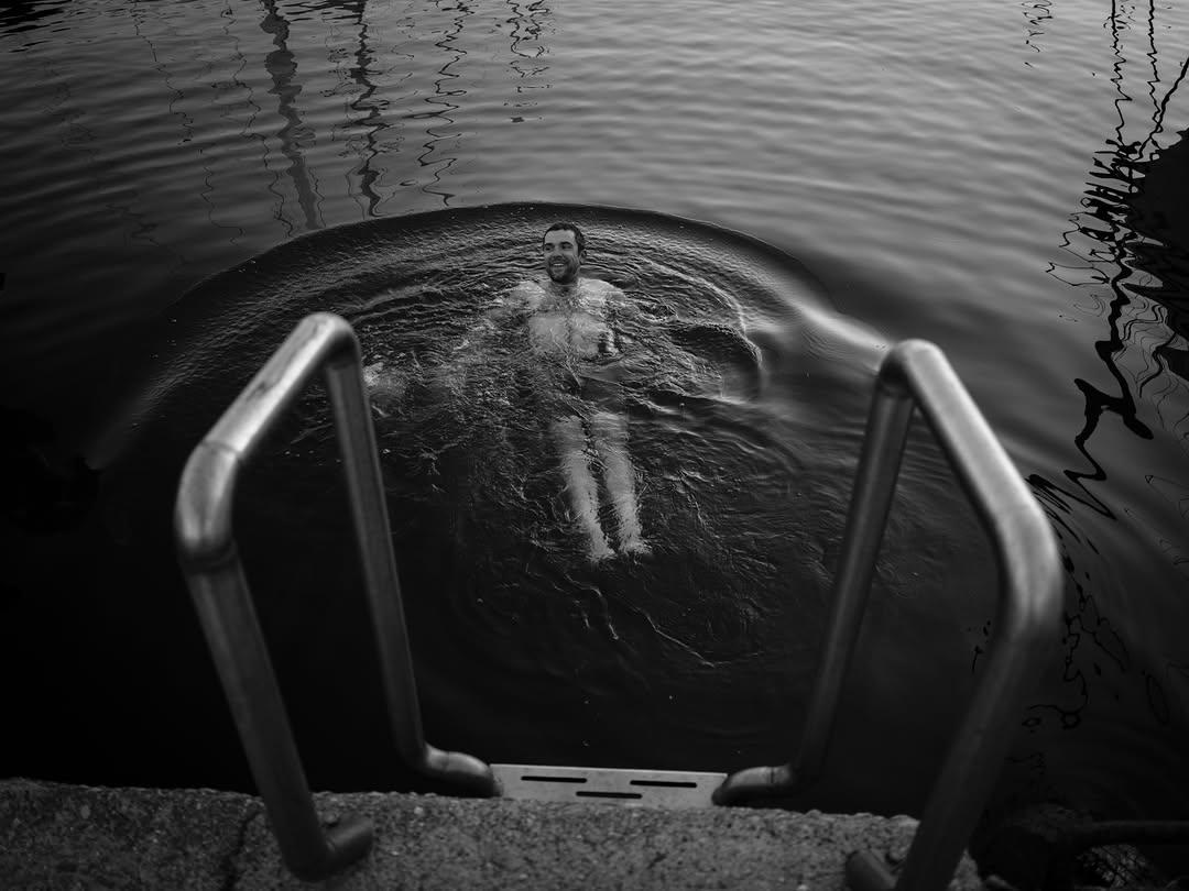 Robin rückenschwimmend im Wasser, Schwarzweißaufnahme. Vor ihm eine Trittleiter ins Wasser.